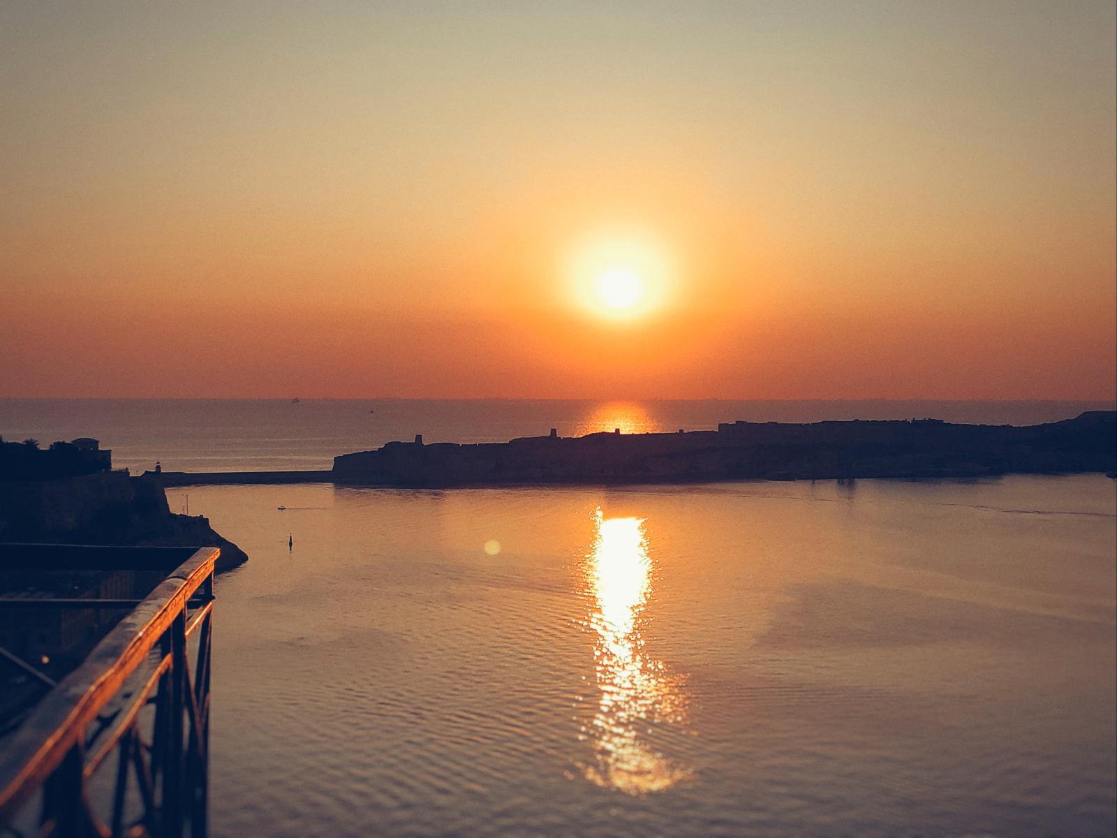 Beeindruckende Aussicht auf den Sonnenuntergang über dem Grand Harbour, Valletta, mit warmen Farbtönen, die sich im Meer spiegeln.