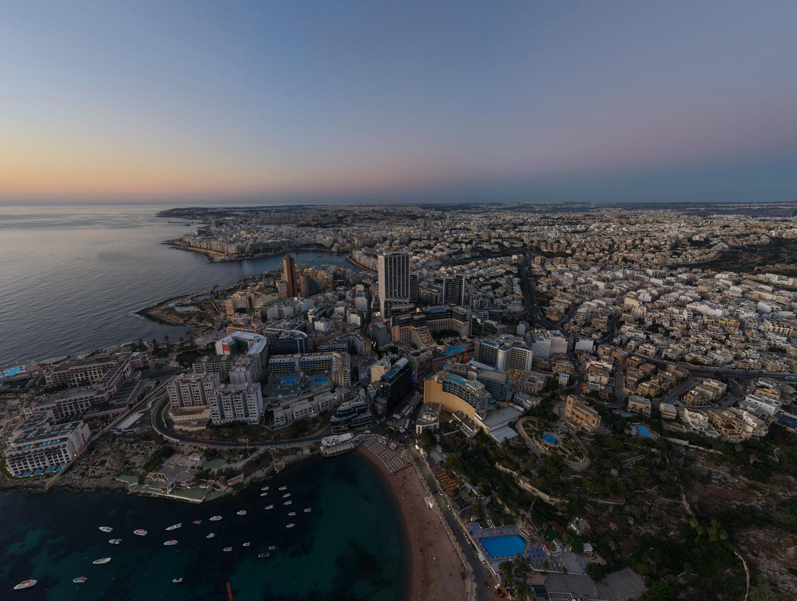 Entdecken Sie eine atemberaubende Luftaufnahme von St. Julian’s, Malta, während die Sonne untergeht und die Küste und das Stadtbild erleuchtet.