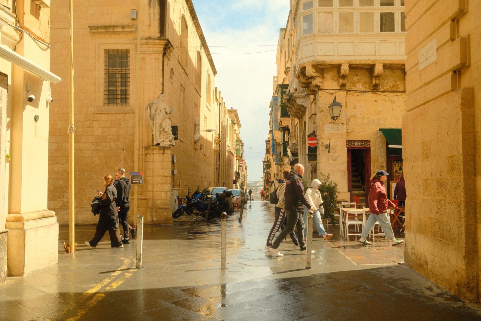 Fußgänger, die entlang einer sonnigen Straße in einer historischen Stadt in Malta gehen, mit traditioneller Architektur aus Kalkstein.
