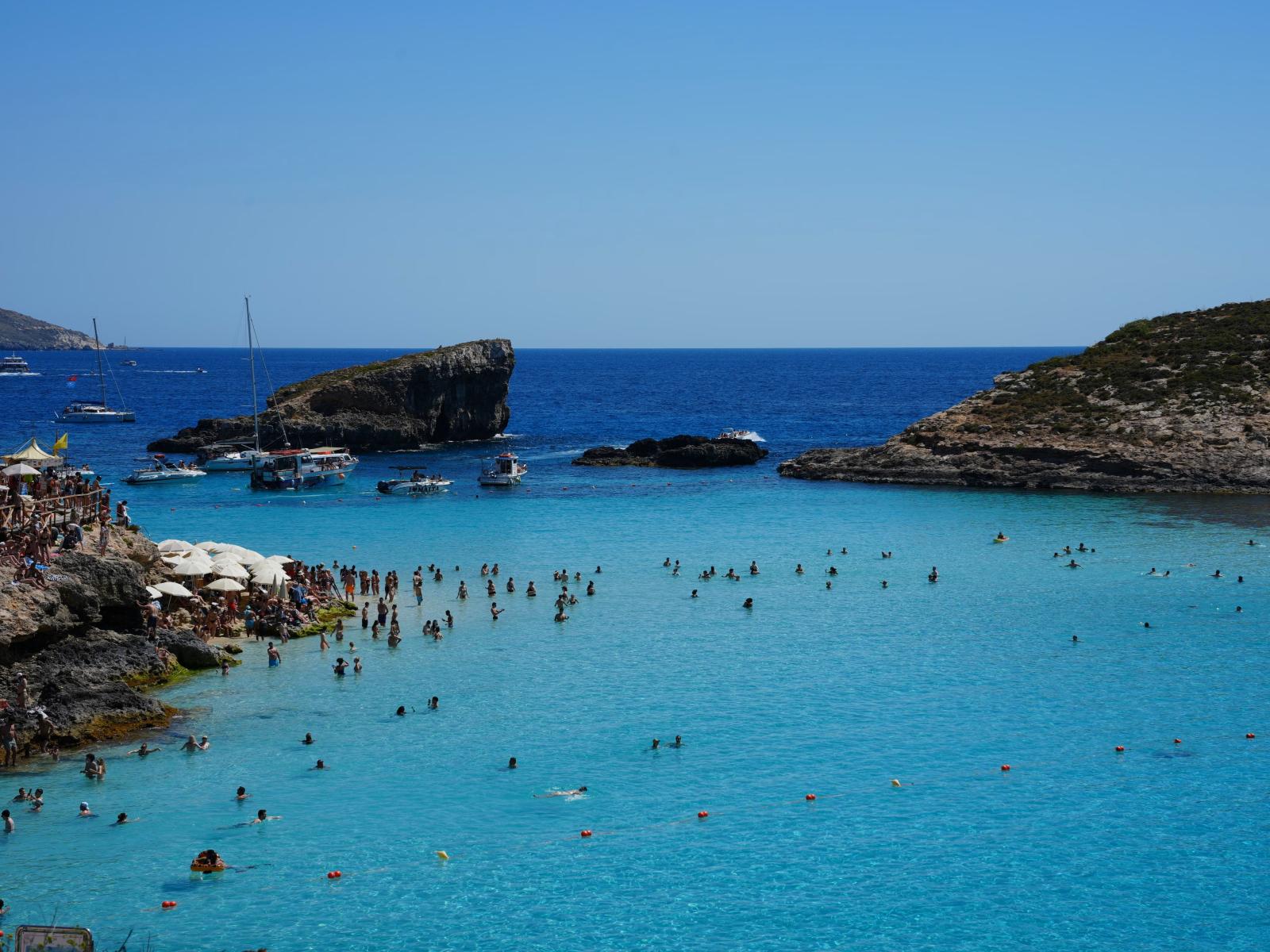 Menschenmengen genießen das klare blaue Wasser der Blauen Lagune auf Malta mit Booten und malerischen Felsen.