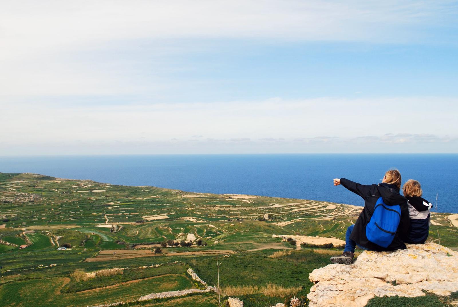 Familie genießt einen Panoramablick auf die Küste Maltas, sitzt auf einem Felsen mit dem Meer und Feldern darunter.