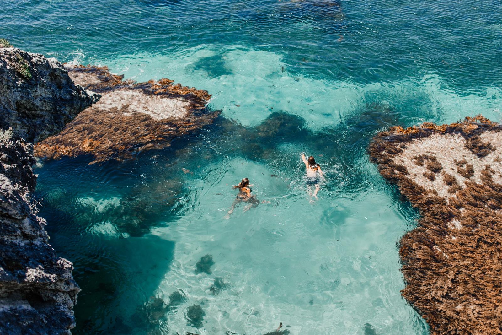 Luftaufnahme von zwei Frauen, die in klarem türkisfarbenem Meer in der Nähe einer felsigen Küste schwimmen.