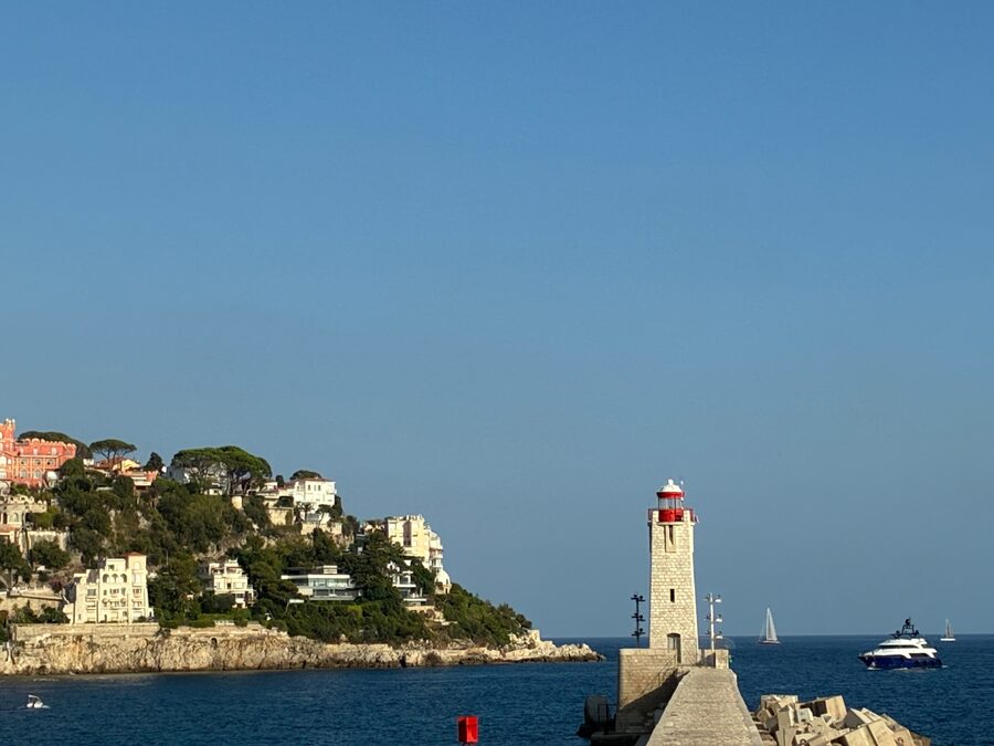 Die Mole in Nizza mit rot-weißem Leuchtturm, im Hintergrund die Küste mit Häusern und ein Boot auf dem Meer